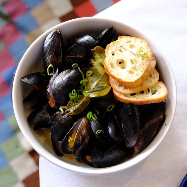 A white bowl of BrewRiver Creole Kitchen's mussels with broth, garnished with scallions and toasted bread.