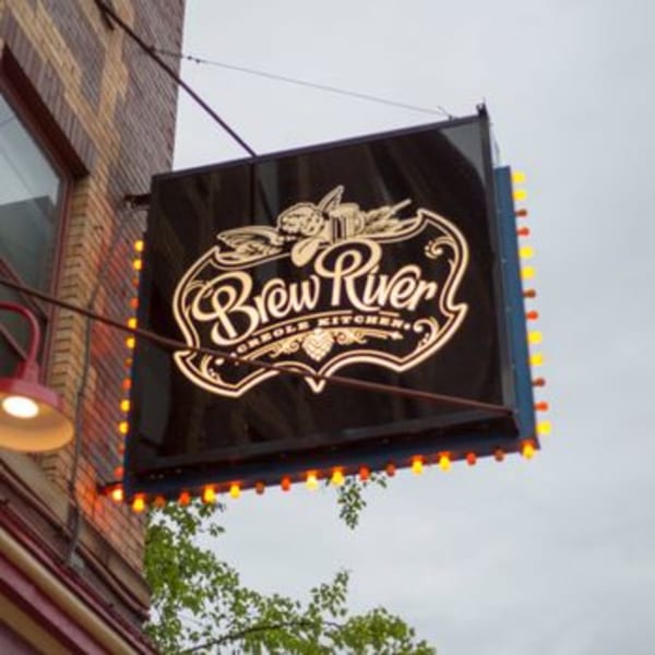 BrewRiver Creole Kitchen's illuminated black sign with a decorative white logo, hanging from a brick building.