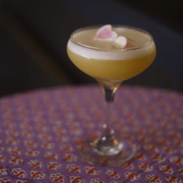 A yellow cocktail with foam and two heart-shaped marshmallows sits on a purple patterned table.