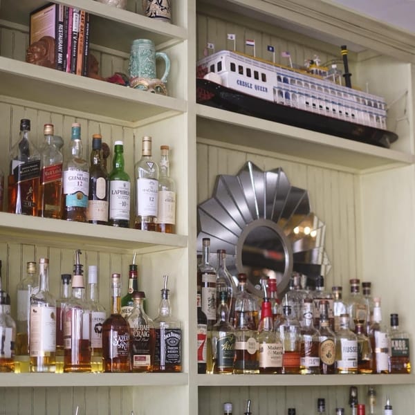 A bar shelf filled with various liquor bottles, books, and a "Creole Queen" steamboat model.