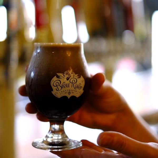 Hands holding a BrewRiver Gastropub branded glass filled with a dark beverage.