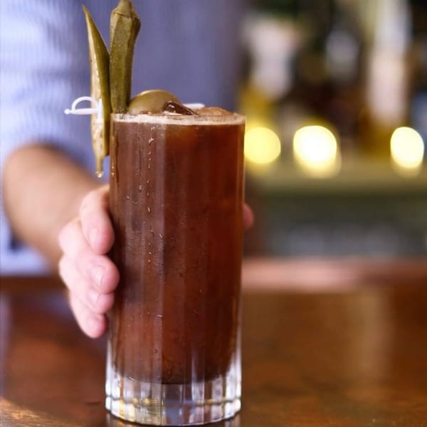 A hand holds a tall glass of a dark creole cocktail garnished with pickled okra and a lime.